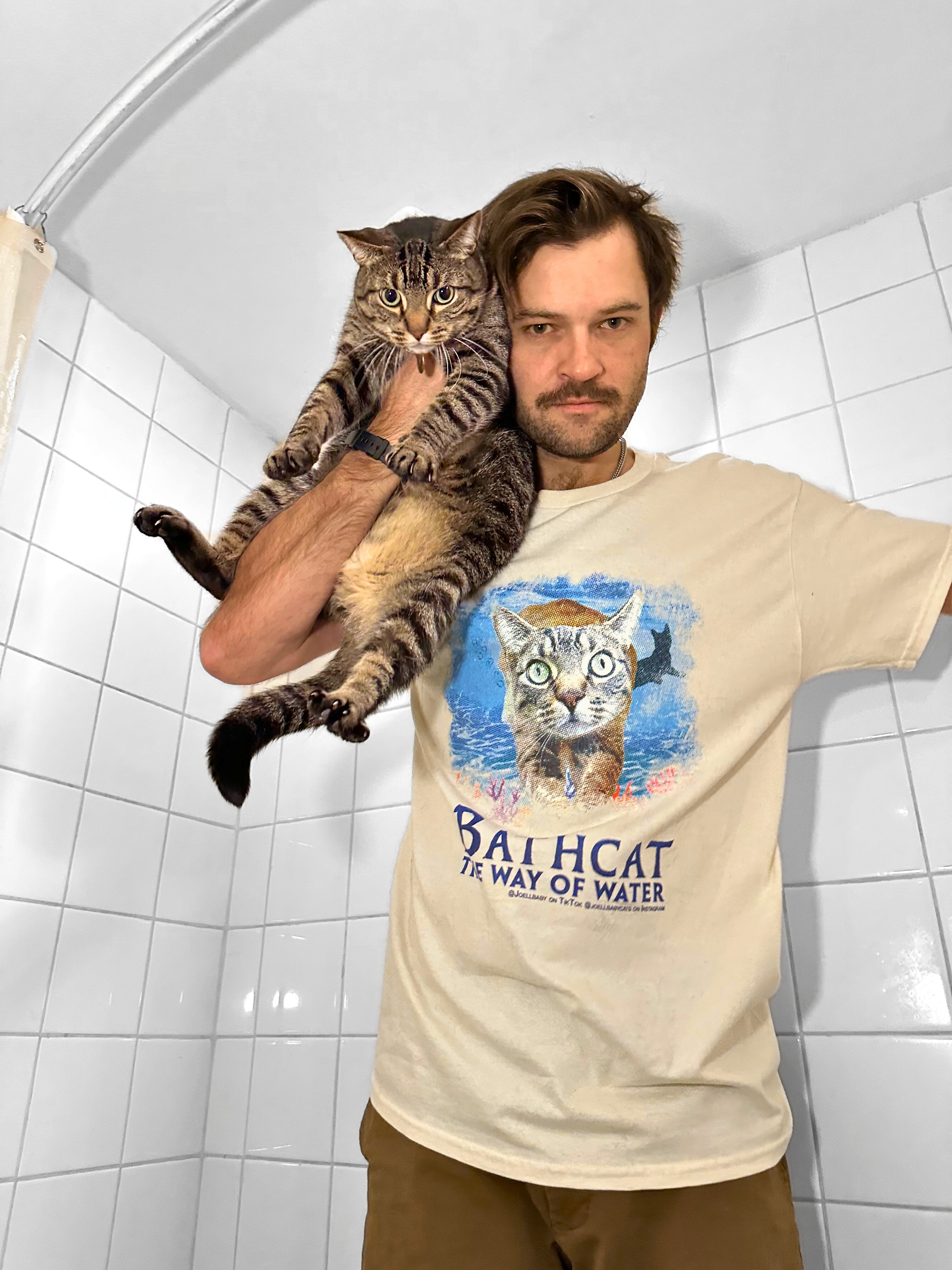 Man holding a cat and wearing a t-shirt with a cat graphic and text in a tiled room.
